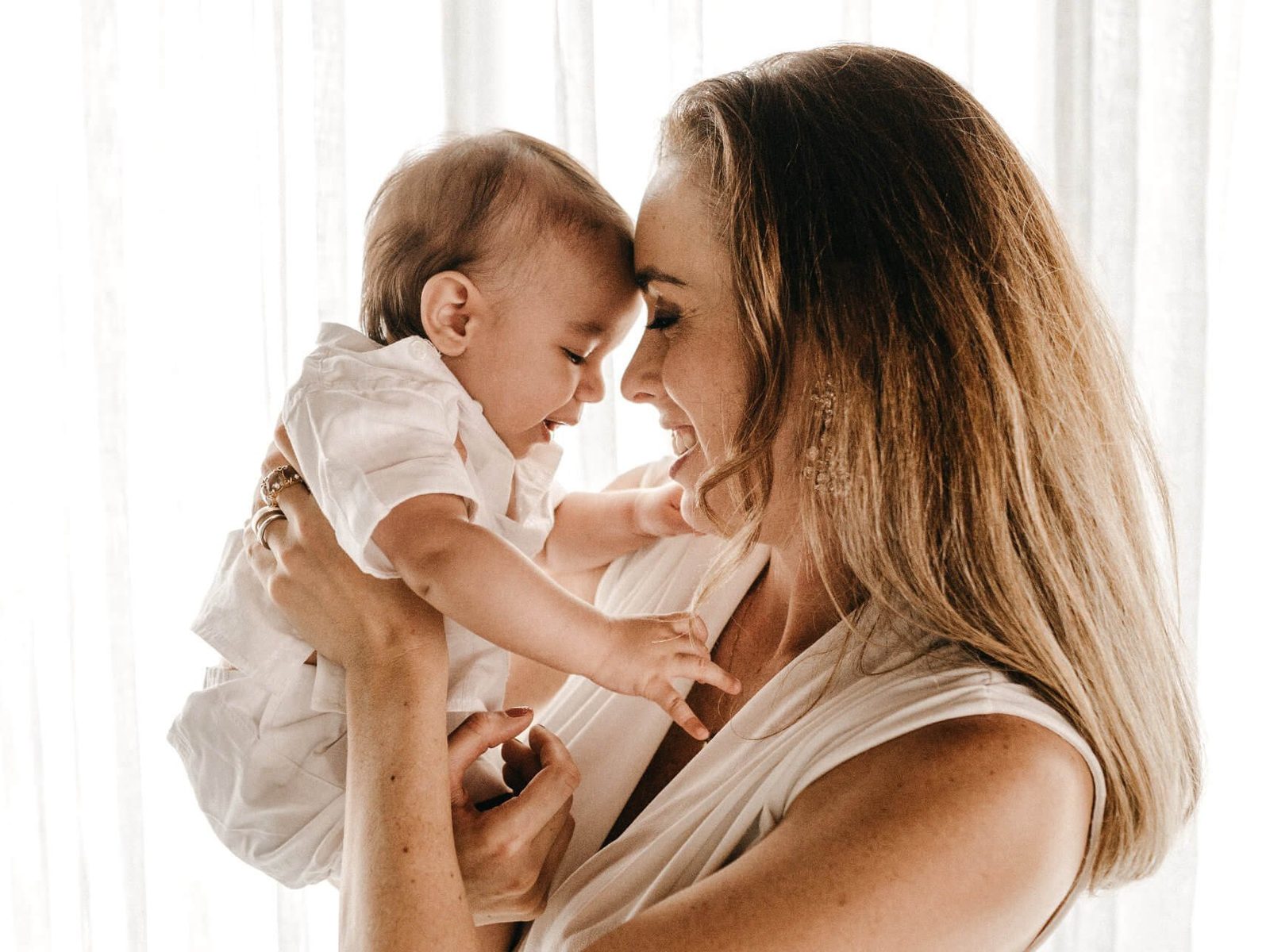 A mother lifting her smiling baby, their foreheads touching, in soft window light